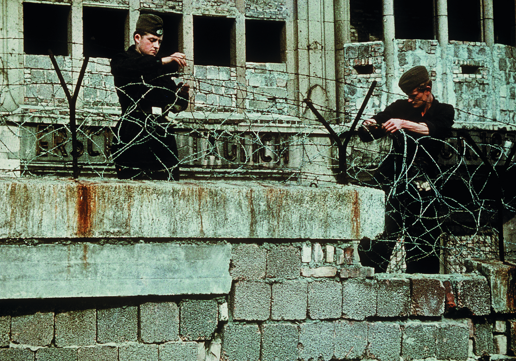 Fotografia. Vista parcial de muro, sobre o qual dois soldados instalam cerca de arame farpado. Ao fundo, vista parcial de prédio em construção.