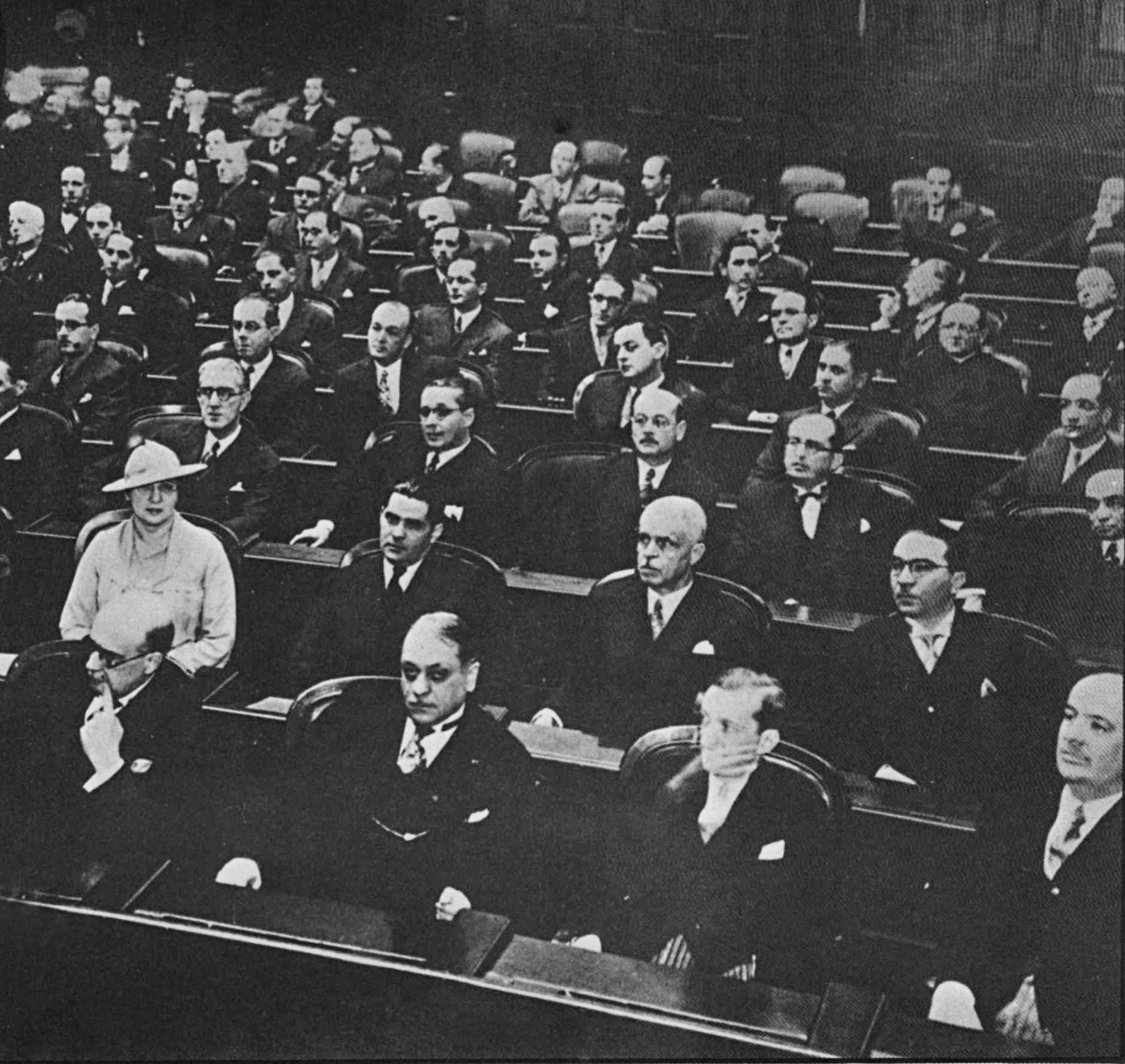 Fotografia em preto e branco. Cerca de dez fileiras de cadeiras escuras com diversos homens sentados. Quase todos eles usam terno preto e camisa branca. Entre eles, na segunda fileira, à esquerda, destaca-se uma única mulher, que usa chapéu e roupas claras.