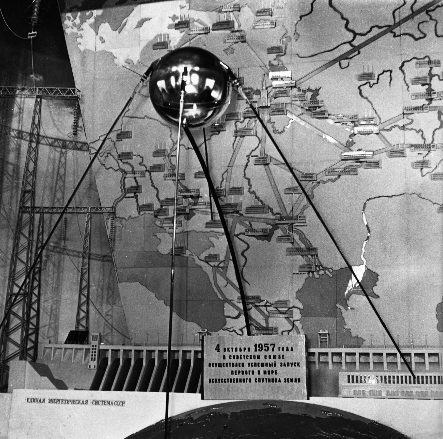 Fotografia em preto e branco. Um objeto em formato esférico e cor metálica está apoiado sobre três suportes de aço, exposto sobre uma mesa. Na parte inferior, um placa com texto escrito em russo, com o ano 1957 em destaque. Ao fundo, grande imagem com ilustração similar a um mapa.
