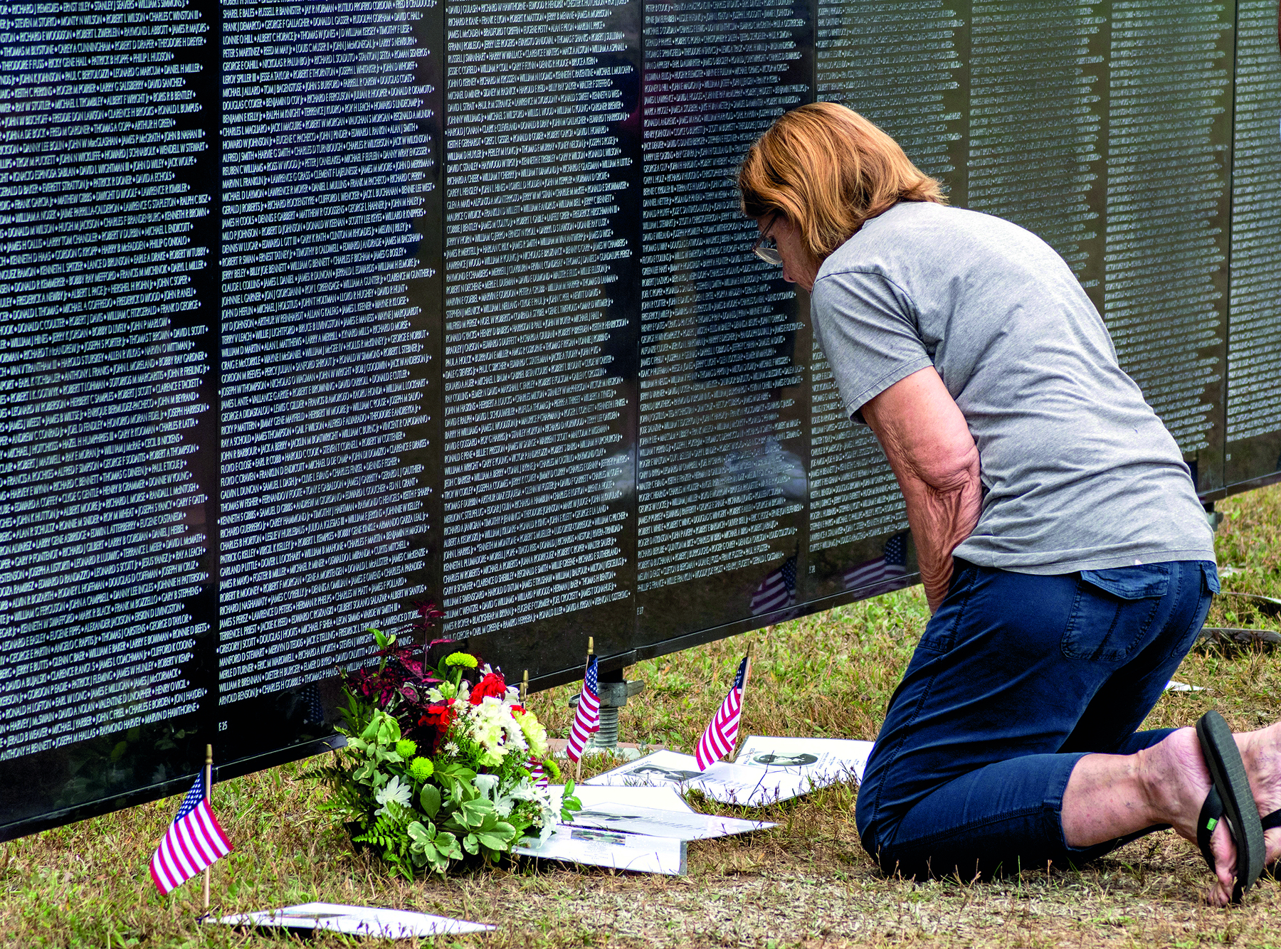 Fotografia. Uma mulher de joelhos diante de uma placa com milhares de nomes escritos. À sua frente, no chão, algumas folhas brancas e três pequenas bandeiras dos Estados Unidos. Entre as bandeiras, um buquê com flores em vermelho, branco e folhas verdes.