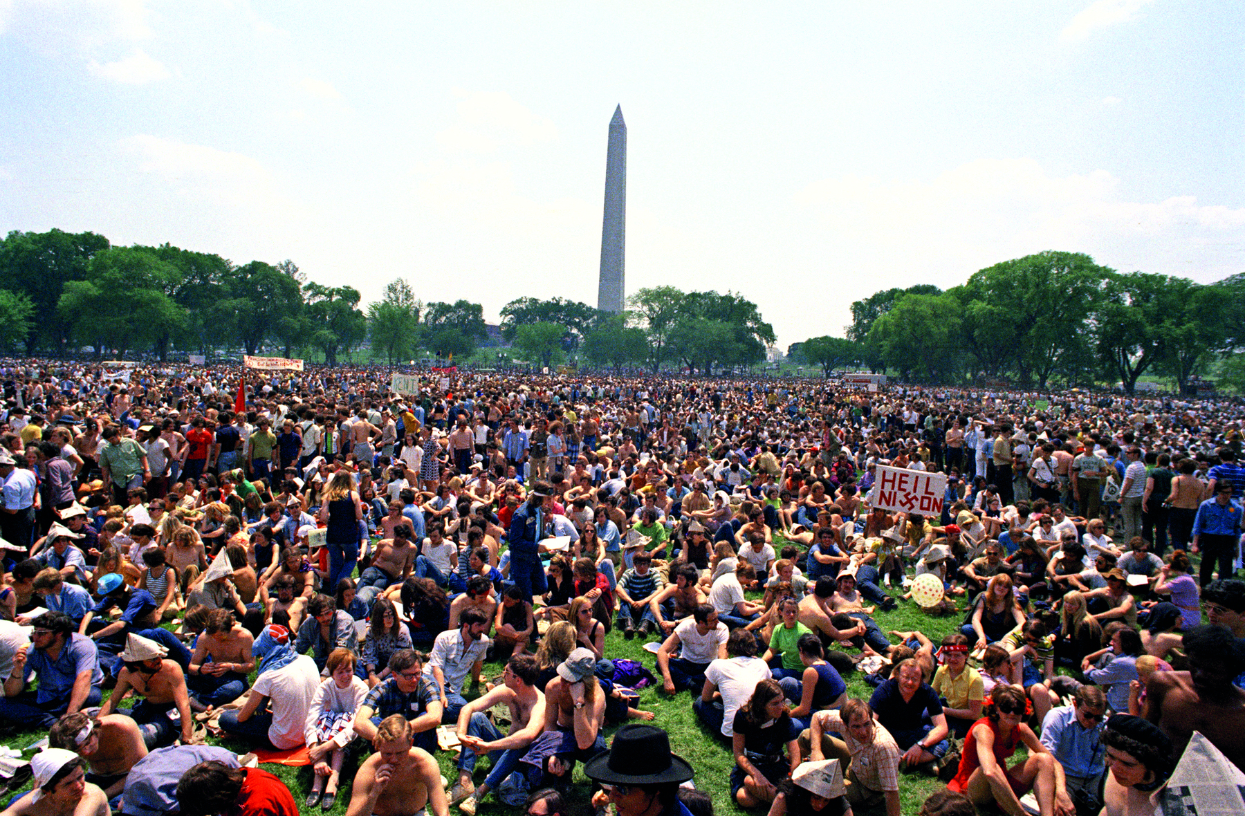 Fotografia. Vista de local aberto com centenas de pessoas sentadas sobre uma grama verde. Ao fundo, muitas árvores e um obelisco. No alto, céu azul-claro e nuvens brancas.