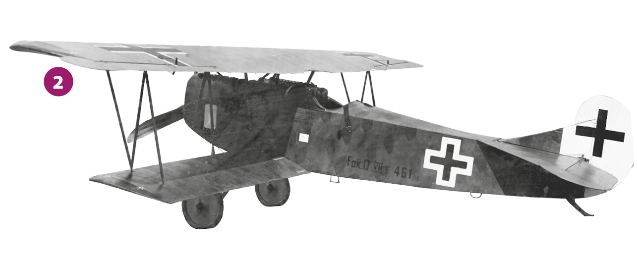 Fotografia 2 em preto e branco. Um avião pequeno, com uma  cruz preta na cauda e outra na lateral. Trem de pouso fixo e hélice na parte frontal