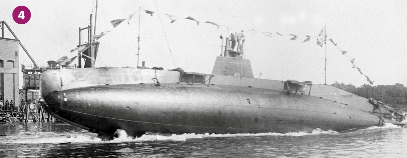 Fotografia 4 em preto e branco. Em local aberto, um submarino emerso sobre a água. Ao fundo, área portuária.