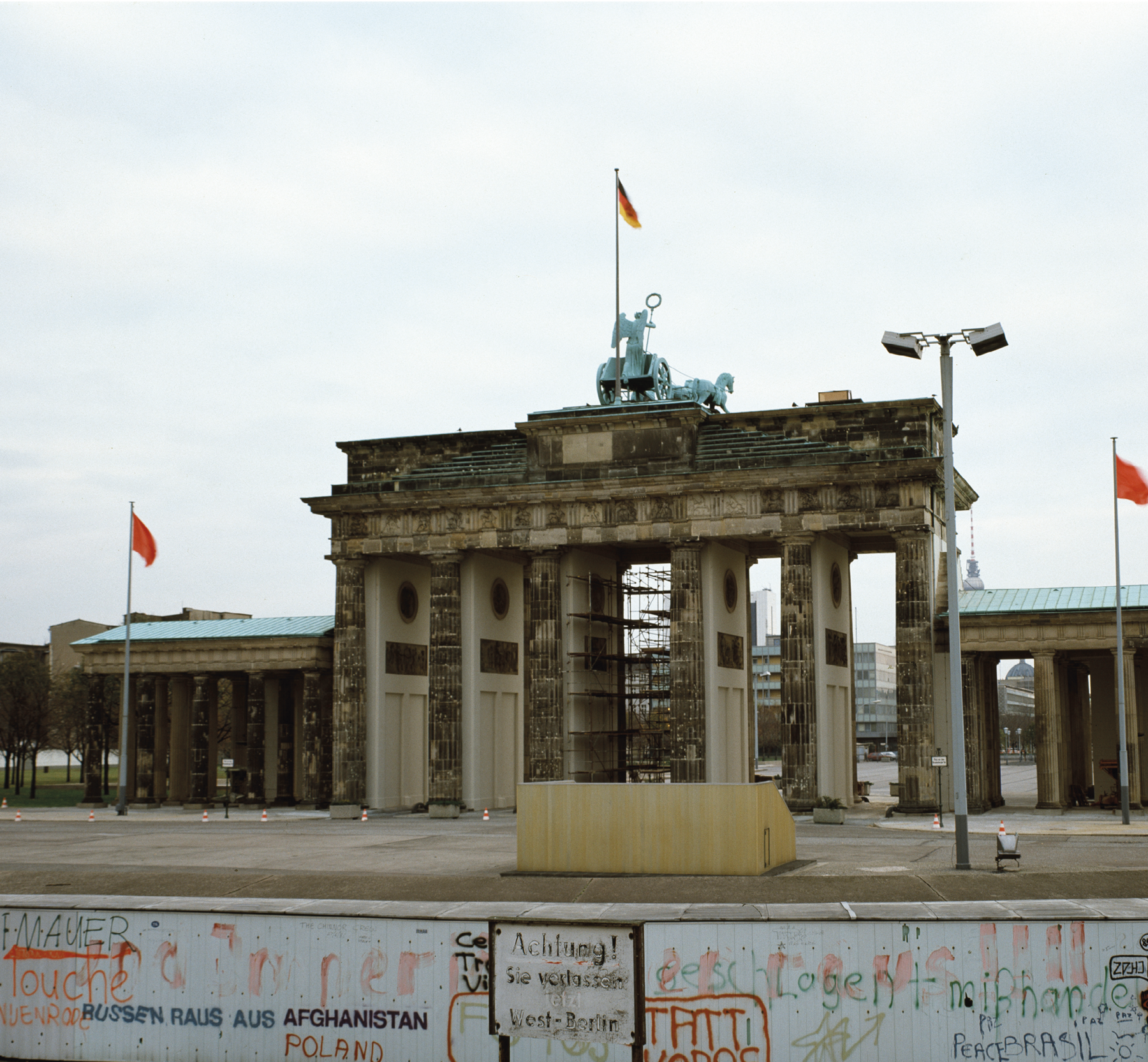 Fotografia. Em lugar aberto, vê-se um muro em primeiro plano, na parte inferior da imagem, cheio de inscrições. Do outro lado do muro, ao fundo, um enorme portão, com seis colunas e cobertura superior, com uma estátua acima e a bandeira da Alemanha. Trata-se do Portão de Brandenburg, na cidade de Berlim. No alto, céu claro com nuvens esparsas.