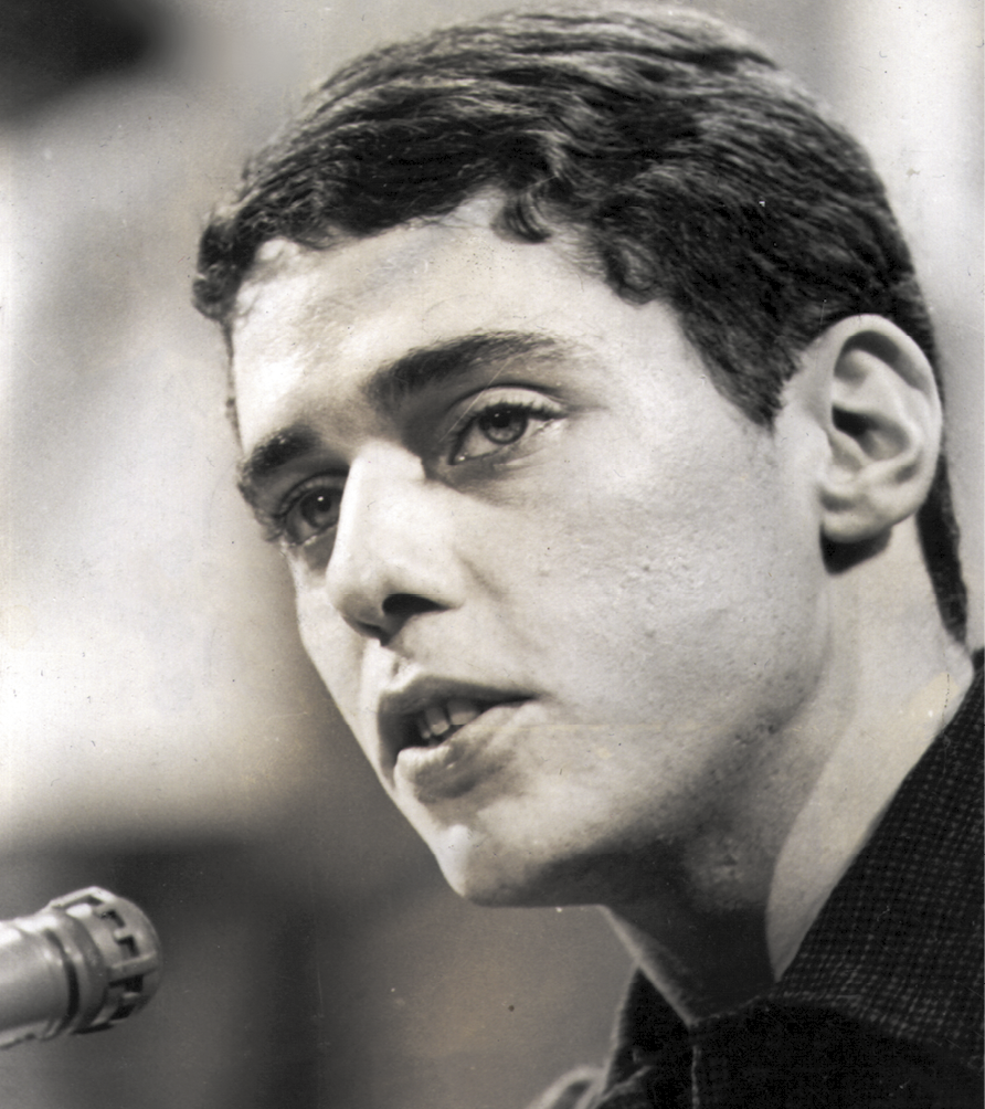 Fotografia em preto e branco. Um homem visto parcialmente, com a cabeça próxima a um microfone, canta, olhando ao longe. Ele tem cabelos escuros, olhos claros e lábios semi-abertos.