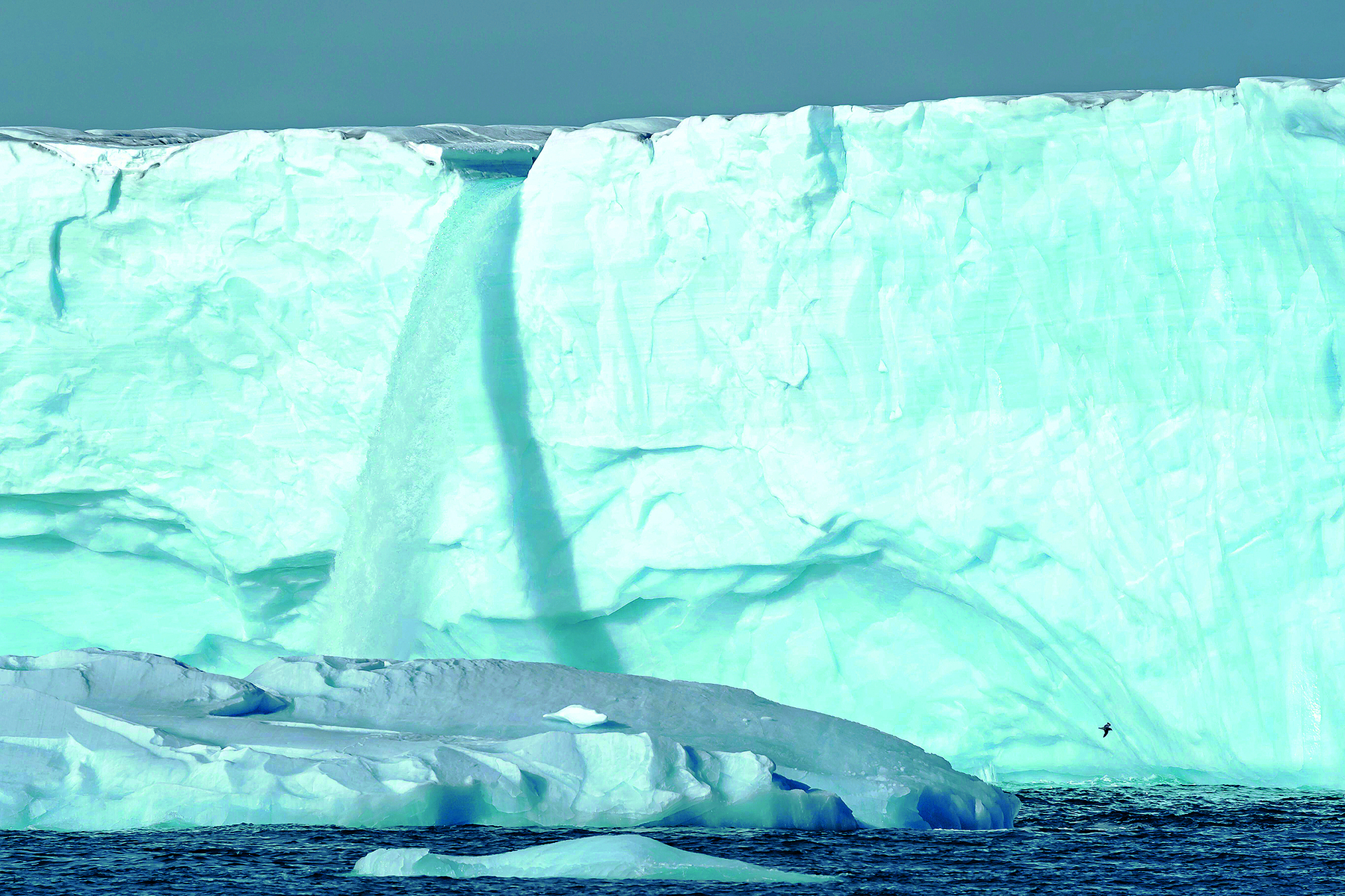 Fotografia. Uma grande geleira em tons de azul-claro, na horizontal, diante do oceano em tons de azul-escuro. No alto, céu em cinza. Ao centro da geleira, uma queda de água que cai sobre o oceano.