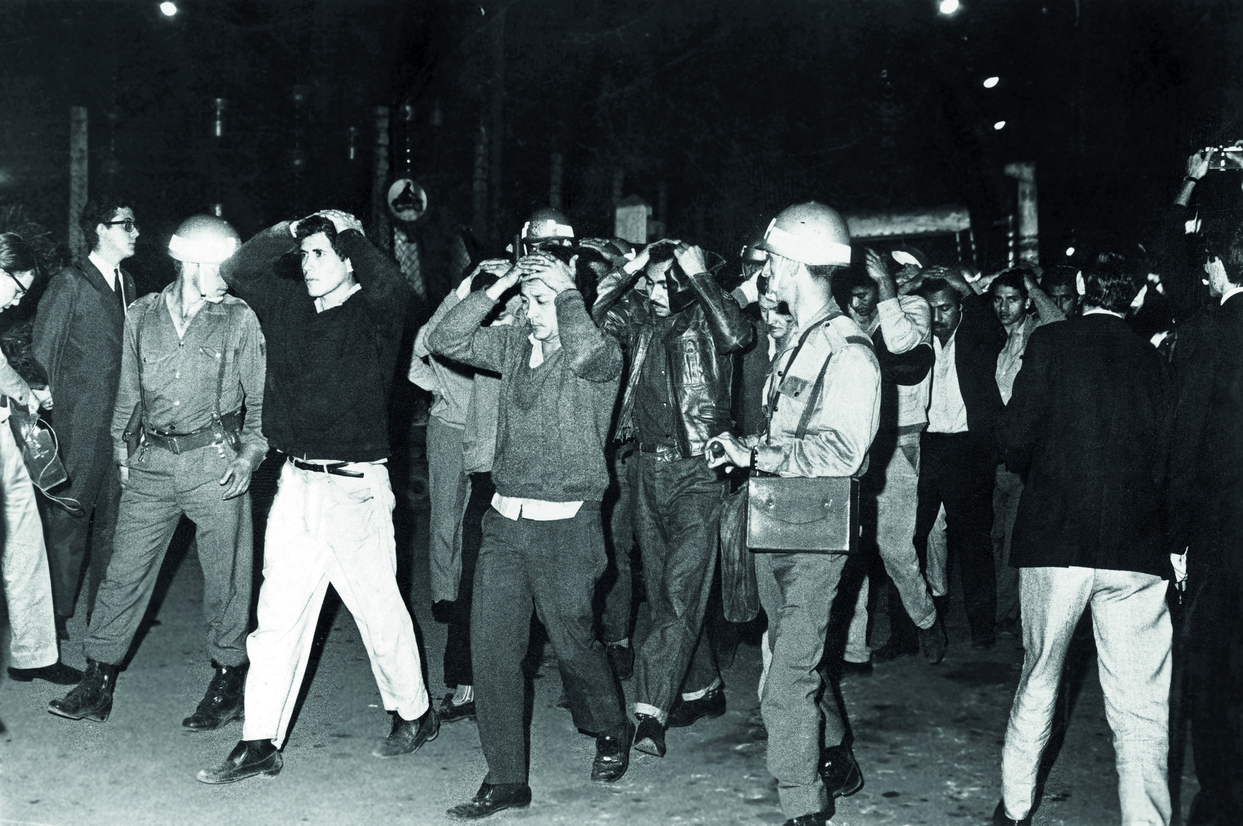 Fotografia em preto e branco. Local aberto, à noite. Em primeiro plano,  muitos homens caminham pela rua com as duas mãos sobre a cabeça, acompanhados de policiais armados. À direita, alguns homens de paletó escuro observam o movimento e fotografam as pessoas.