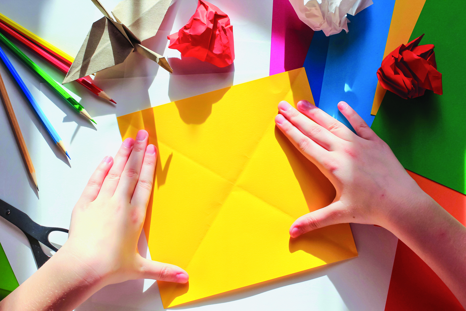 Fotografia. Mãos de pessoa sobre uma folha amarela de formato quadrado, com marcas de dobra nas diagonais. No canto superior esquerdo, lápis de diferentes cores. Na parte superior algumas bolas de papel e uma ave feita com dobraduras.  À direita, folhas de diferentes cores.  No canto inferior esquerdo uma tesoura preta,