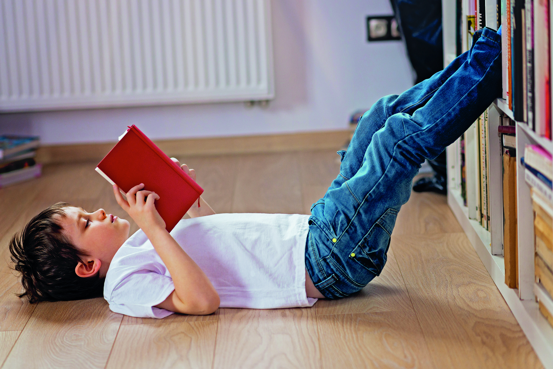 Fotografia. Menino de camiseta branca e calça jeans, deitado no chão com as pernas para cima apoiadas na estante, lendo um livro de capa vermelha.