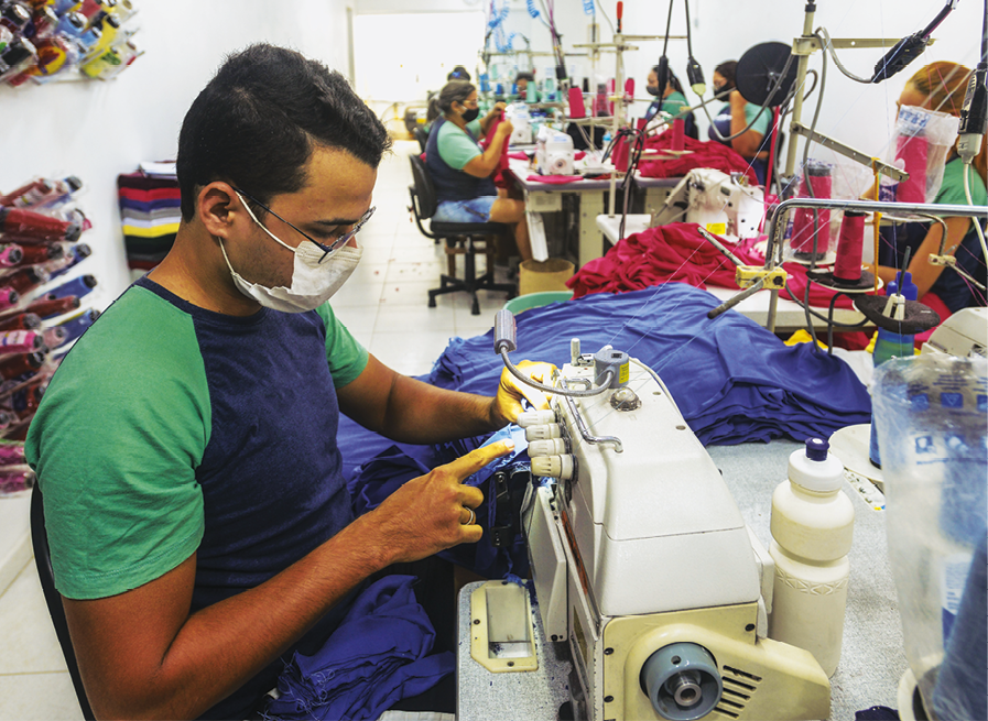 Fotografia. Homem de pele branca e cabelo preto, óculos e máscara de proteção, sentado manuseando uma máquina de costurar. Ao fundo outras pessoas manuseando máquinas de costura.