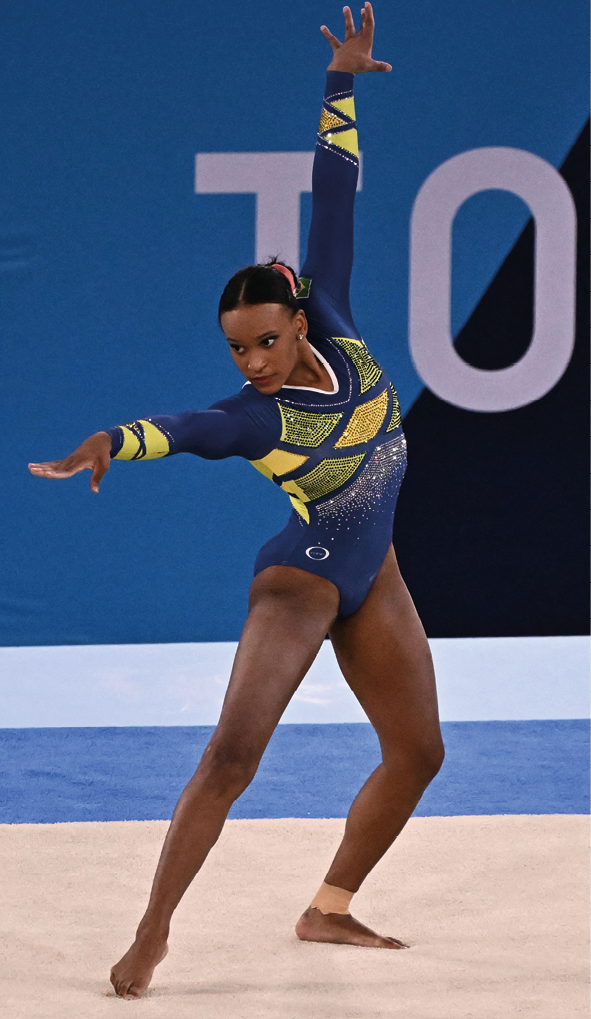 Fotografia. Ginasta brasileira Rebeca Andrade, em uma competição de ginástica de solo. Rebeca é uma mulher negra, está usando um uniforme de competição que é parecido com um maiô de mangas longas, azul com figuras geométricas em verde e amarelo. Está em uma posição de apresentação com um braço para cima e outro para frente.