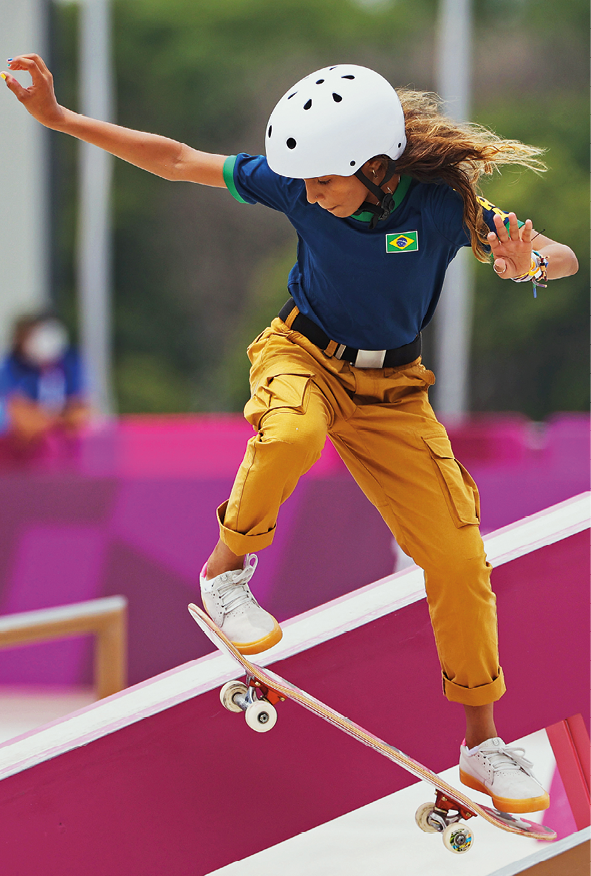 Fotografia. Skatista brasileira chamada Rayssa Leal fazendo uma manobra com o Skate. Menina de cabelos longos e castanhos, está com capacete branco, vestindo camiseta azul marinho com a bandeira do Brasil e uma calça amarela.