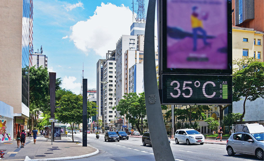 Fotografia. Paisagem urbana da Avenida Paulista, em São Paulo. Em primeiro plano, um poste com um telão e um visor de temperatura indicando 35 graus Celsius. Ao fundo uma rua movimentada com muitos carros e algumas pessoas na calçada. Há também algumas árvores e muitos prédios nos dois lados da rua.