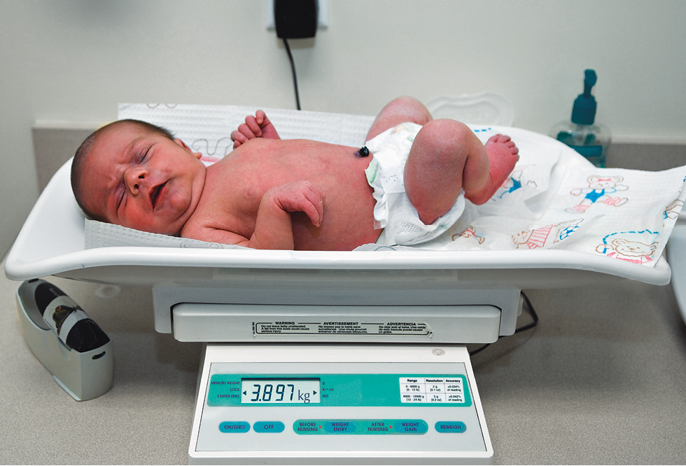 Fotografia. Bebê branco, de fralda, deitado em cima de balança digital branca com detalhes em verde, com visor indicando 3 vírgula 897 quilogramas.