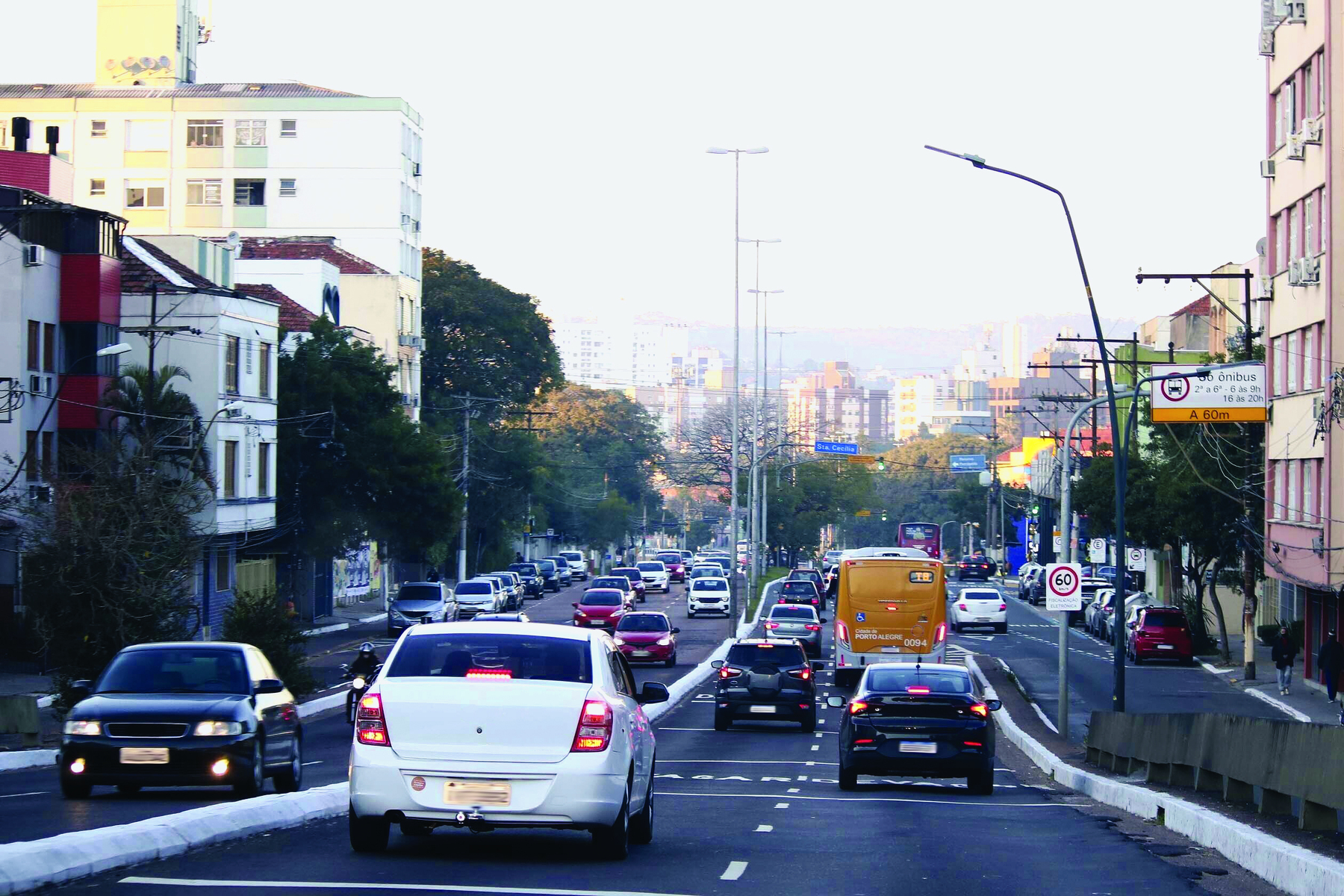 Fotografia. Vista da cidade de Porto Alegre durante o dia com destaque para uma avenida movimentada com muitos carros e ônibus. Ao lado direito é possível identificar alguns prédios pequenos. Ao fundo, algumas árvores, indicando, uma área verde e mais longe muitos prédios que parecem ser mais altos.