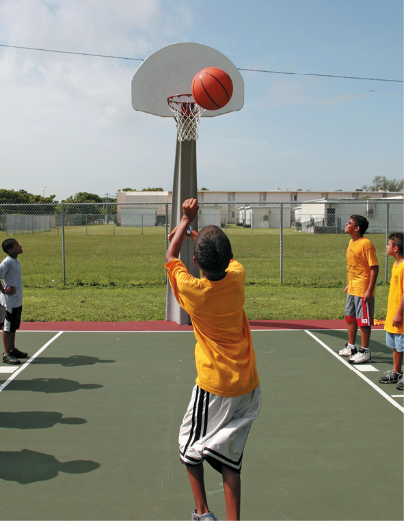 Fotografia. Menino de camiseta amarela, bermuda branca, na quadra arremessando uma bola na cesta de basquete. Em cada lado da cesta os outros jogadores observam o arremesso.