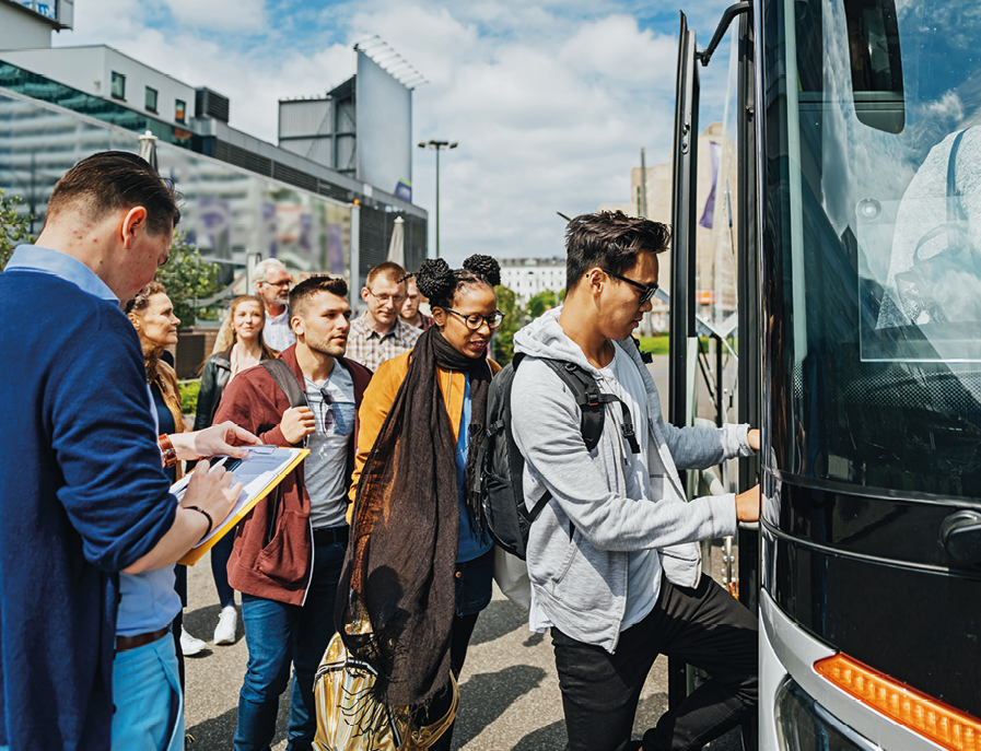 Fotografia. Fila de pessoas entrando em um ônibus. Do lado esquerdo tem um homem escrevendo em uma prancheta.