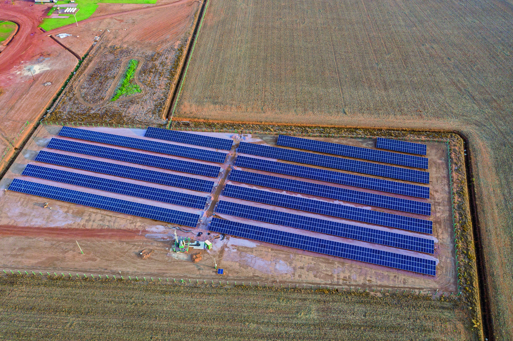Fotografia. Vista do alto de fileiras de painéis de captação de energia solar em chão de terra. O dia está ensolarado.