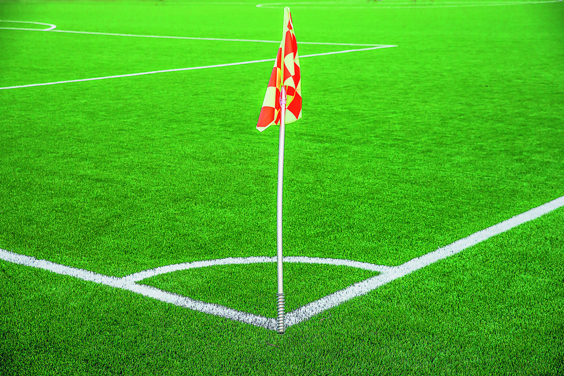 Fotografia. Parte de um campo de futebol, com destaque para um de seus 4 cantos. Na imagem, há uma bandeira  vermelha e branca no encontro da linha lateral com a linha de fundo.