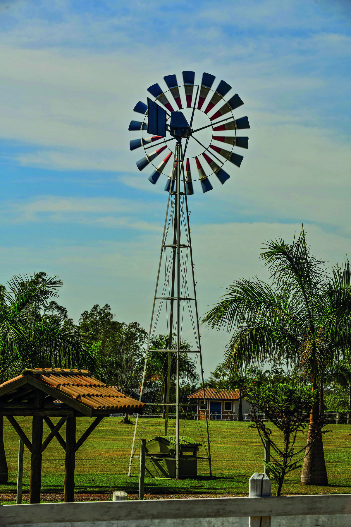 Fotografia. Vista frontal de moinho de vento formado por uma torre de metal e hastes conectadas a um eixo central. O moinho está em um gramado e o dia está ensolarado com nuvens.
