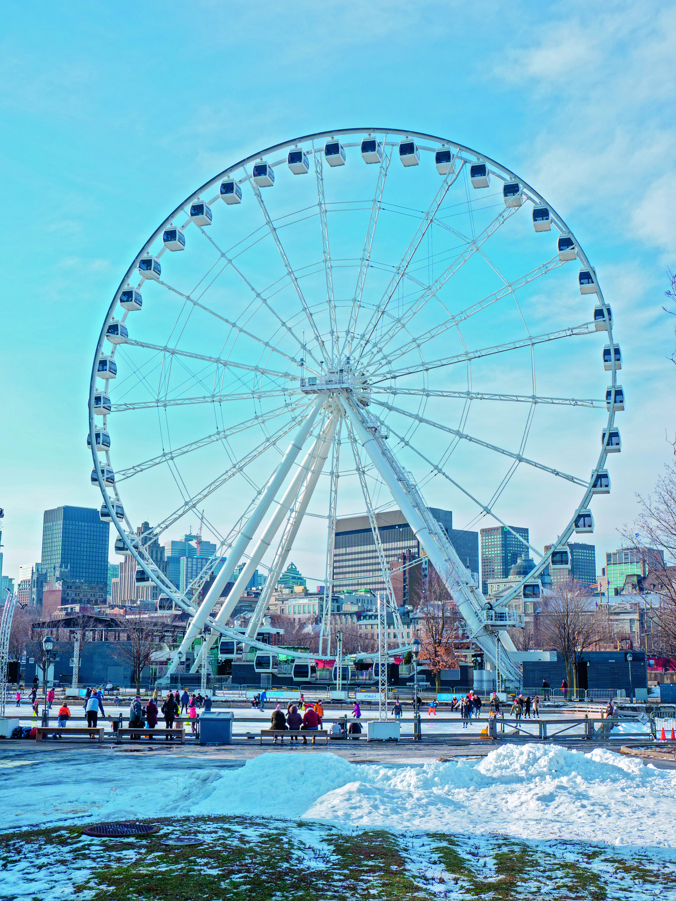 Fotografia. Vista frontal de uma roda-gigante localizada na cidade de Montreal, Canadá, em 2020. É possível perceber seu formato circular e cabines em volta dela. Há pessoas olhando para a roda-gigante. O chão contém neve. Ao fundo da fotografia há prédios da cidade.