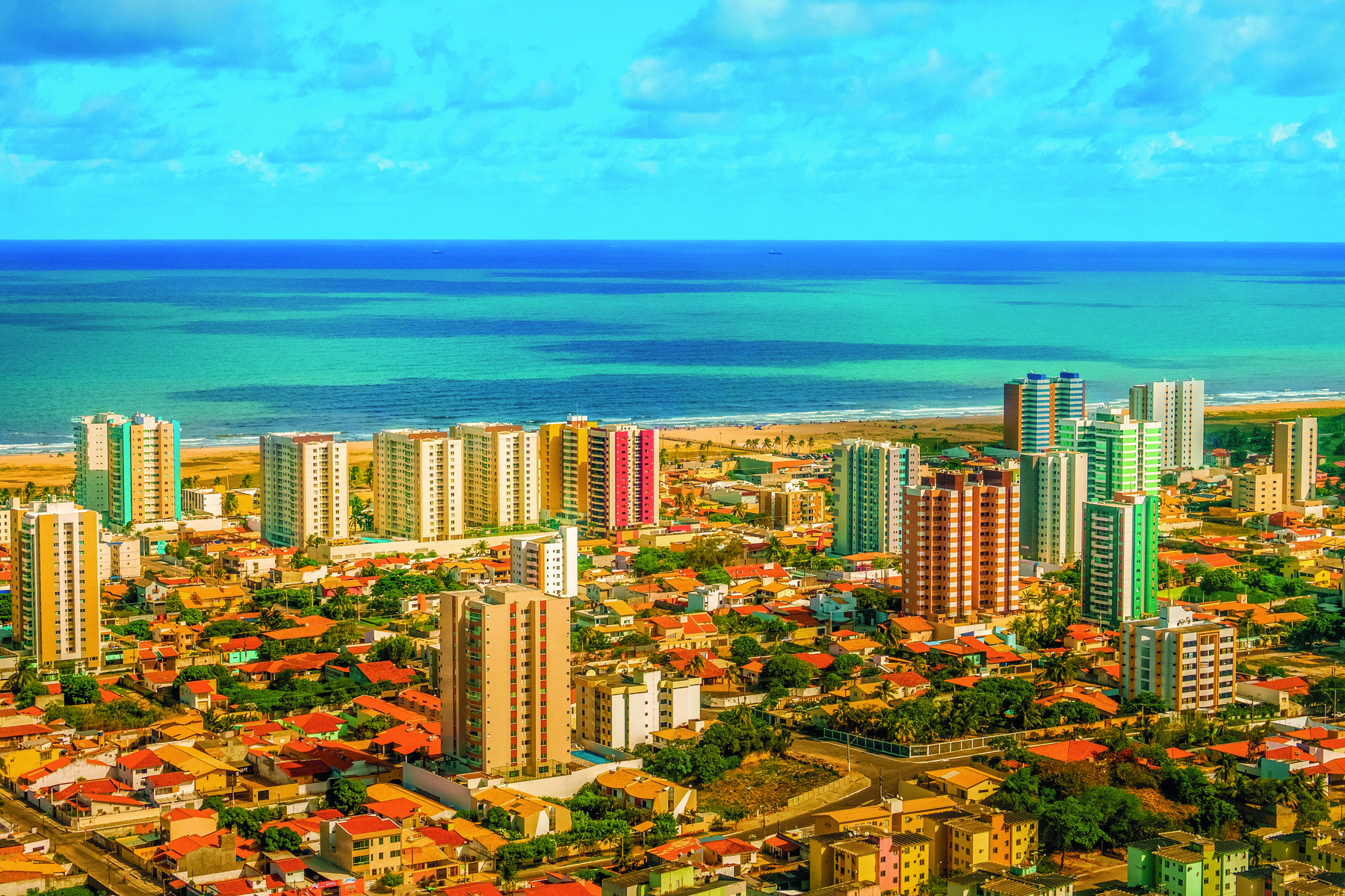 Fotografia. Vista aérea de cidade com casas e prédios ao fundo, o mar está azul. Dia ensolarado com céu azul e poucas nuvens.