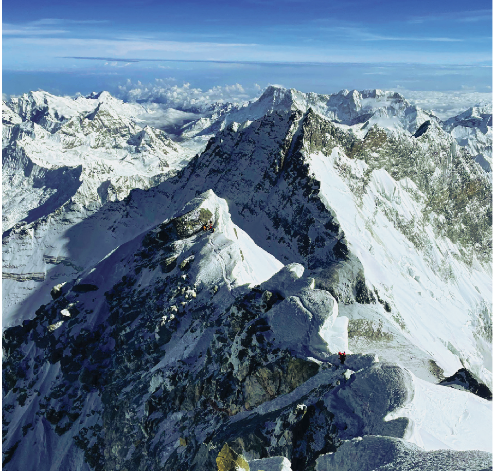 Fotografia. Vista do Monte Everest, em que é possível identificar picos do monte cobertos de neve. O céu está azul com nuvens brancas.