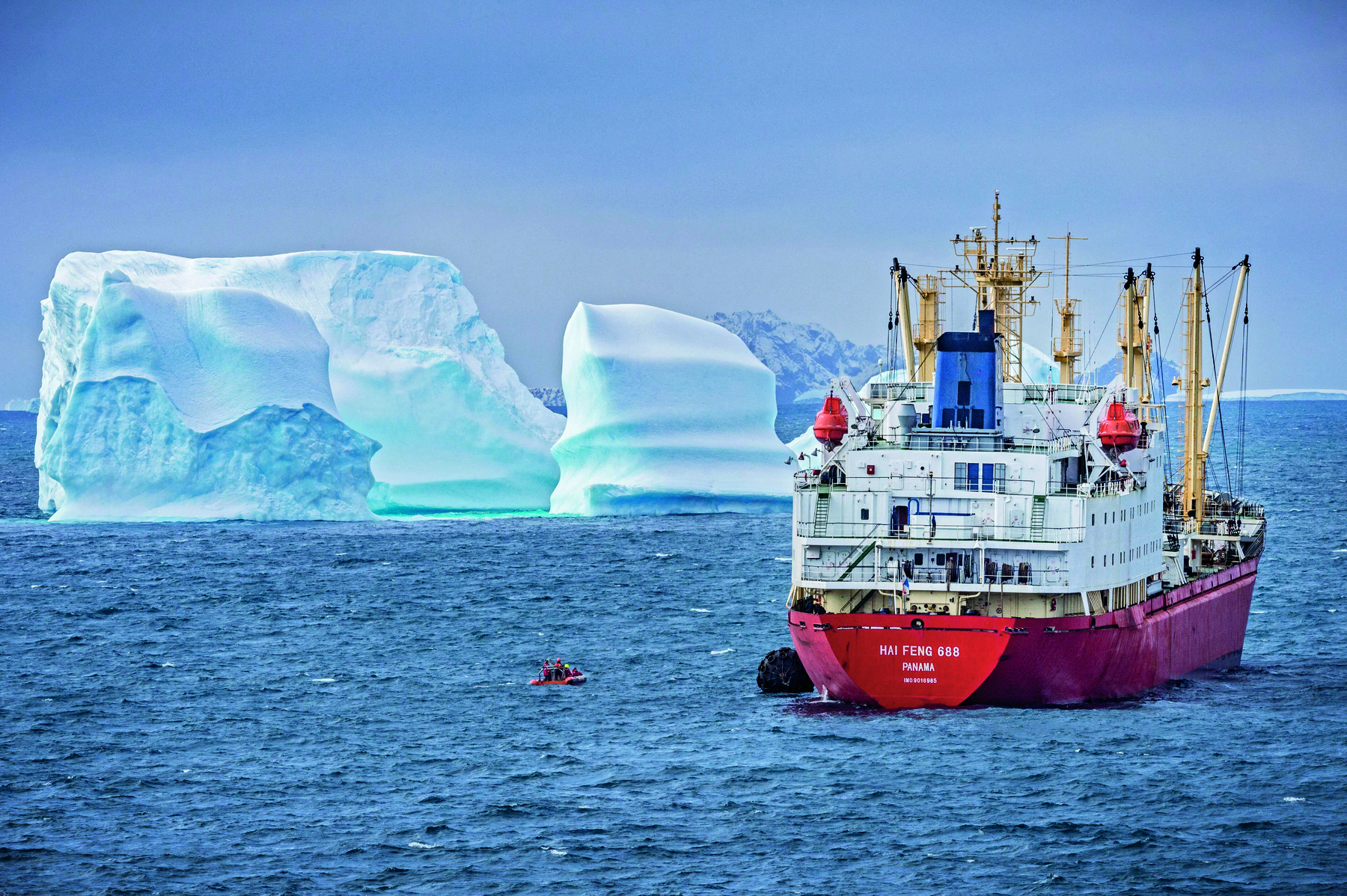 Fotografia. Navio de pesca navegando pelo mar da Antártida. Ele está localizado do lado direito da fotografia. O casco do navio é vermelho. A ponte de comando e as demais instalações são brancas. Ao lado do navio, no mar, há um bote com duas pessoas. Ao fundo da fotografia, lado esquerdo, há icebergs.