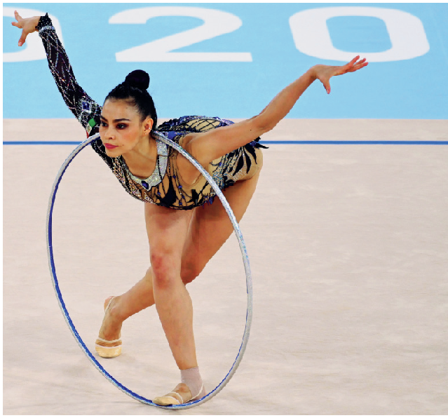 Fotografia. Atleta de ginástica rítmica de cabelo preto e roupa colorida está com os braços para o lado e o tronco abaixado para frente, a cabeça está dentro de um bambolê, apoiado em seu pescoço.