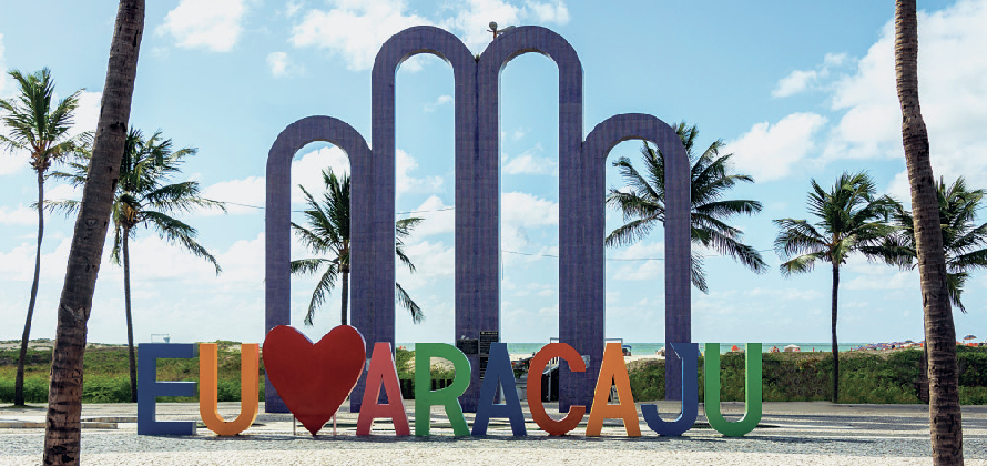 Fotografia. Escultura formada por 4 arcos grandes, um ao lado do outro. Os dois centrais são mais altos que os demais. À frente dos arcos, um letreiro escrito em letras maiúsculas Eu, símbolo de coração, Aracaju.