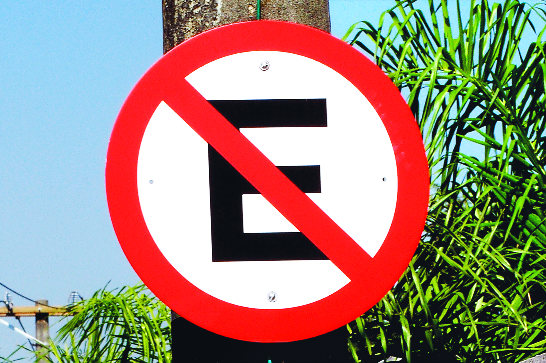 Fotografia. Placa de sinalização de trânsito proibindo estacionar. Placa circular com fundo branco e borda vermelha com a letra E no centro, uma linha diagonal vermelha está sobre a letra E.