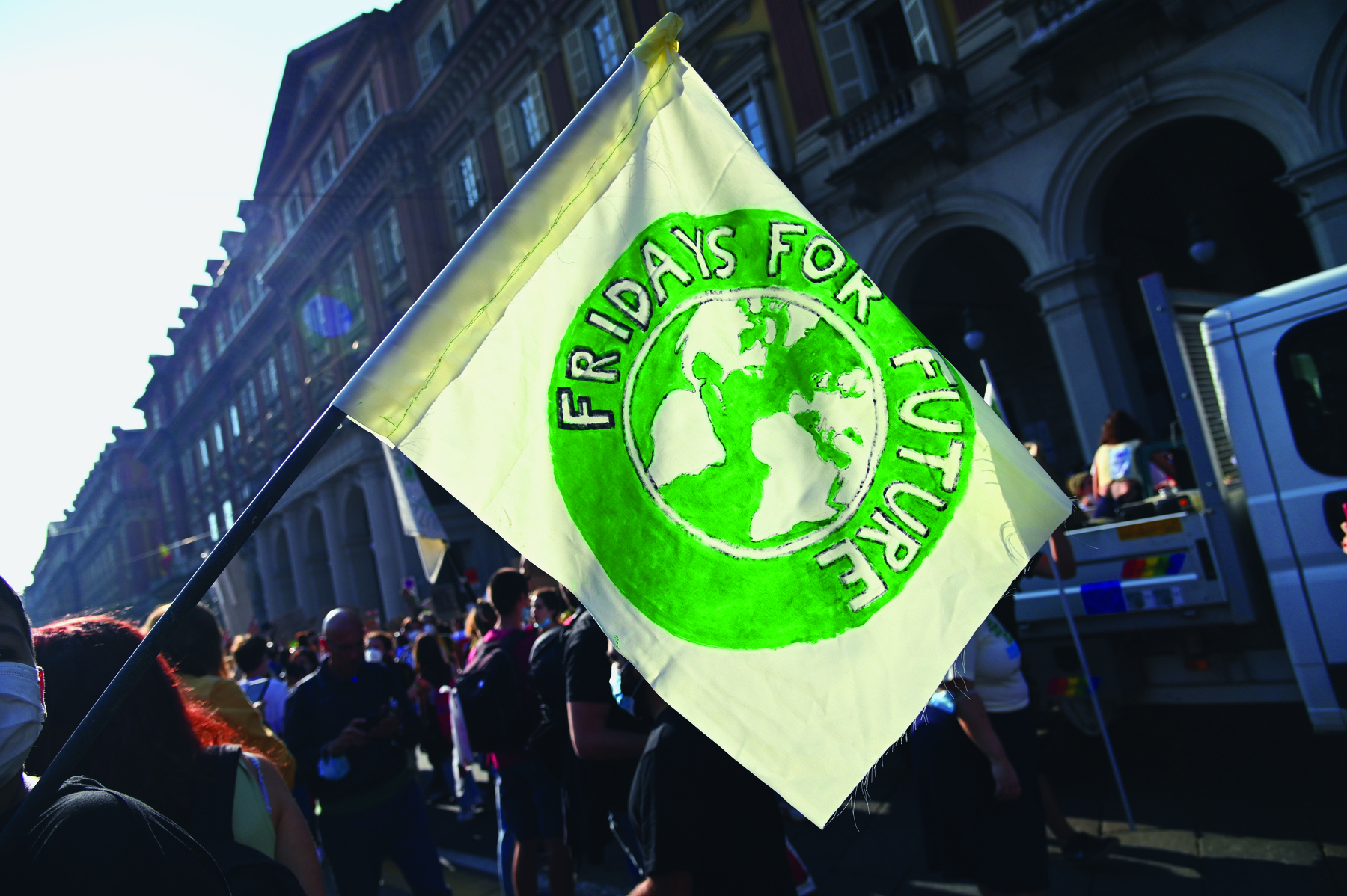 Fotografia. Aglomeração de pessoas em frente a uma brande construção. Uma pessoa segura uma bandeira branca com círculo verde escrito ao redor: FRIDAYS FOR FUTURE. No centro, há uma ilustração do planeta Terra com os continentes em branco.