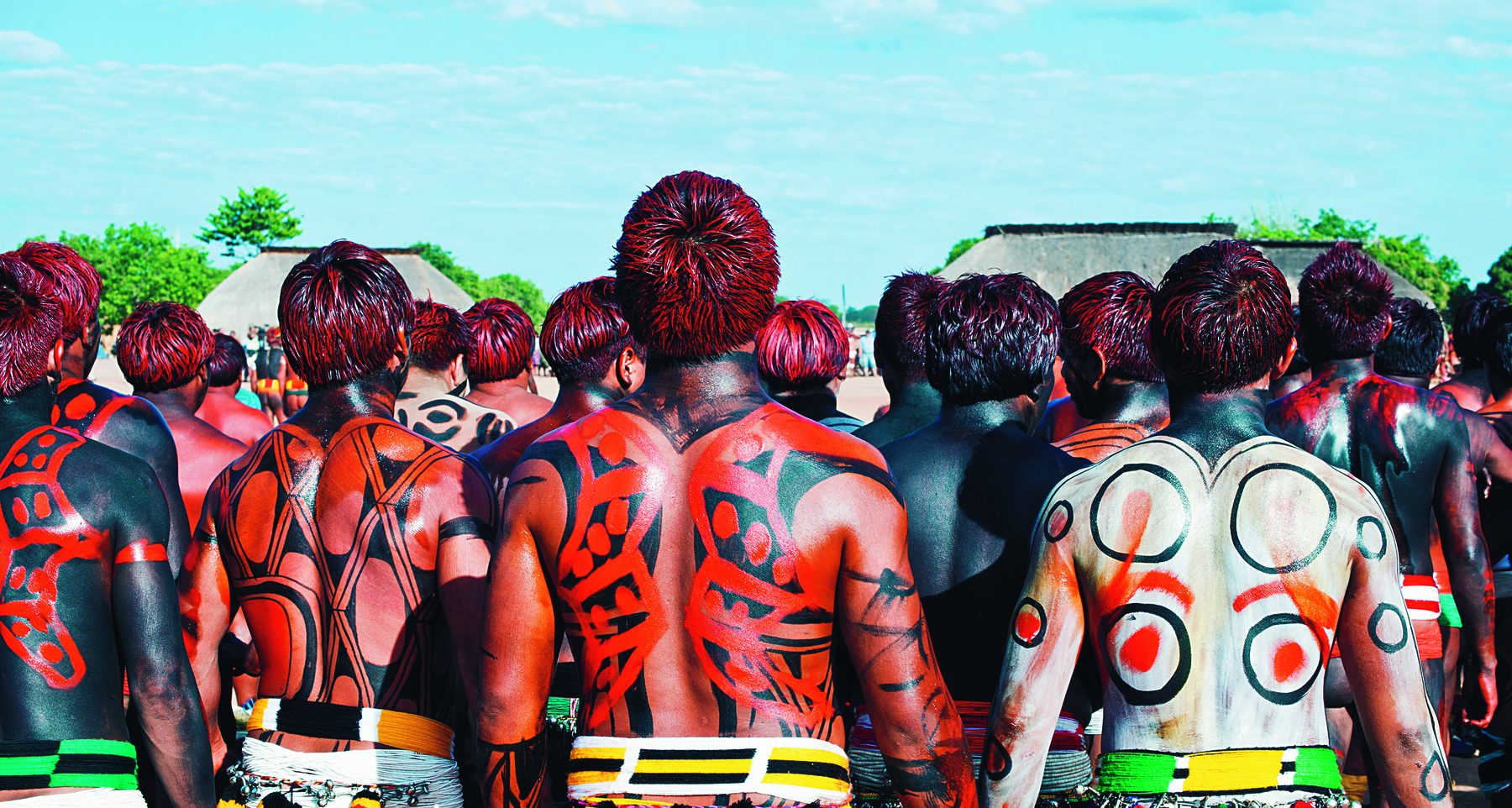Fotografia. Vista de trás de pessoas com os corpos pintados com formas geométricas na cores vermelho, preto e branco.