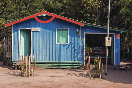 Fotografia. Vista frontal de uma casa quilombola colorida em azul. É possível identificar uma porta aberta, uma janela retangular e uma área aberta como se fosse garagem. O terreno da casa é chão de terra. Há destaque para o ângulo formato no telhado que lembra um triângulo obtusângulo.