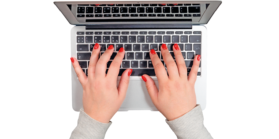 Fotografia. Vista superior de mãos com unhas pintadas com esmalte vermelho mexendo nas teclas de um notebook aberto.