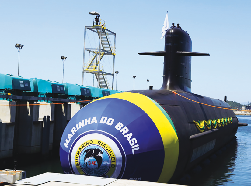 Fotografia. Vista frontal de um submarino flutuando na água. Há um logo da Marinha do Brasil na frente.