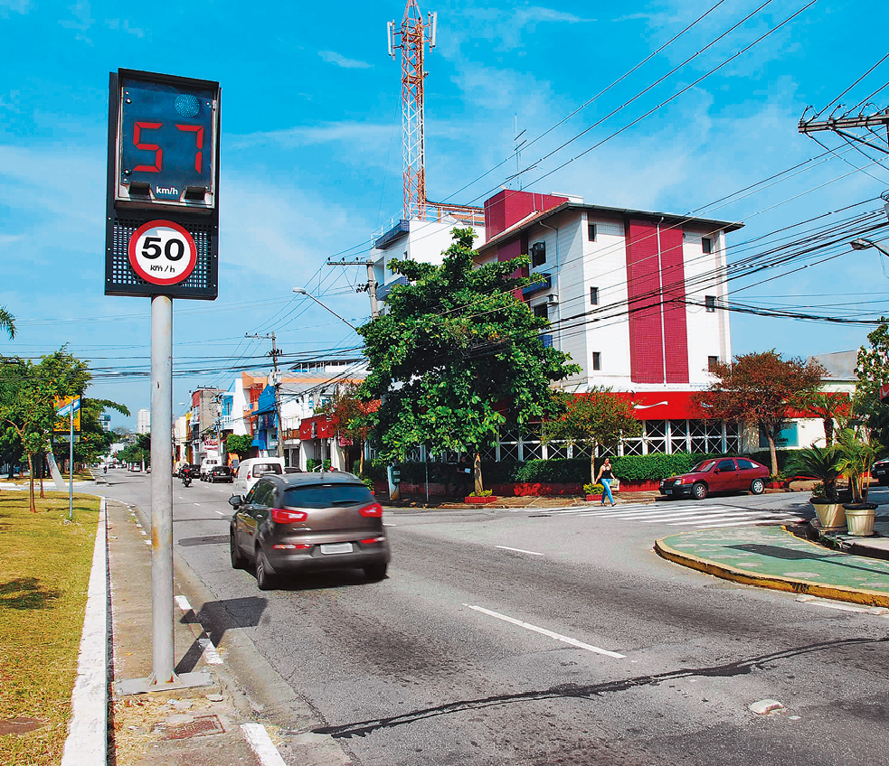 Fotografia. À esquerda, visor de radar indicando a medida de de velocidade de 57 quilômetros por hora. Abaixo, placa de trânsito redonda indicando a velocidade de 50 quilômetros por hora como a máxima permitida. Aparece um carro que acabou de passar pelo radar e ao fundo, construção e mais carros.