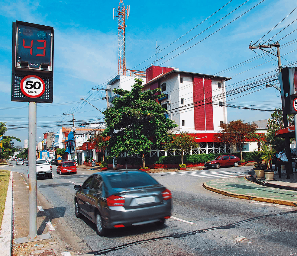 Fotografia. À esquerda, visor de radar indicando a medida de de velocidade de 43 quilômetros por hora. Abaixo, placa de trânsito redonda indicando a velocidade de 50 quilômetros por hora como a máxima permitida. Aparece um carro que acabou de passar pelo radar e ao fundo, construção e mais carros.