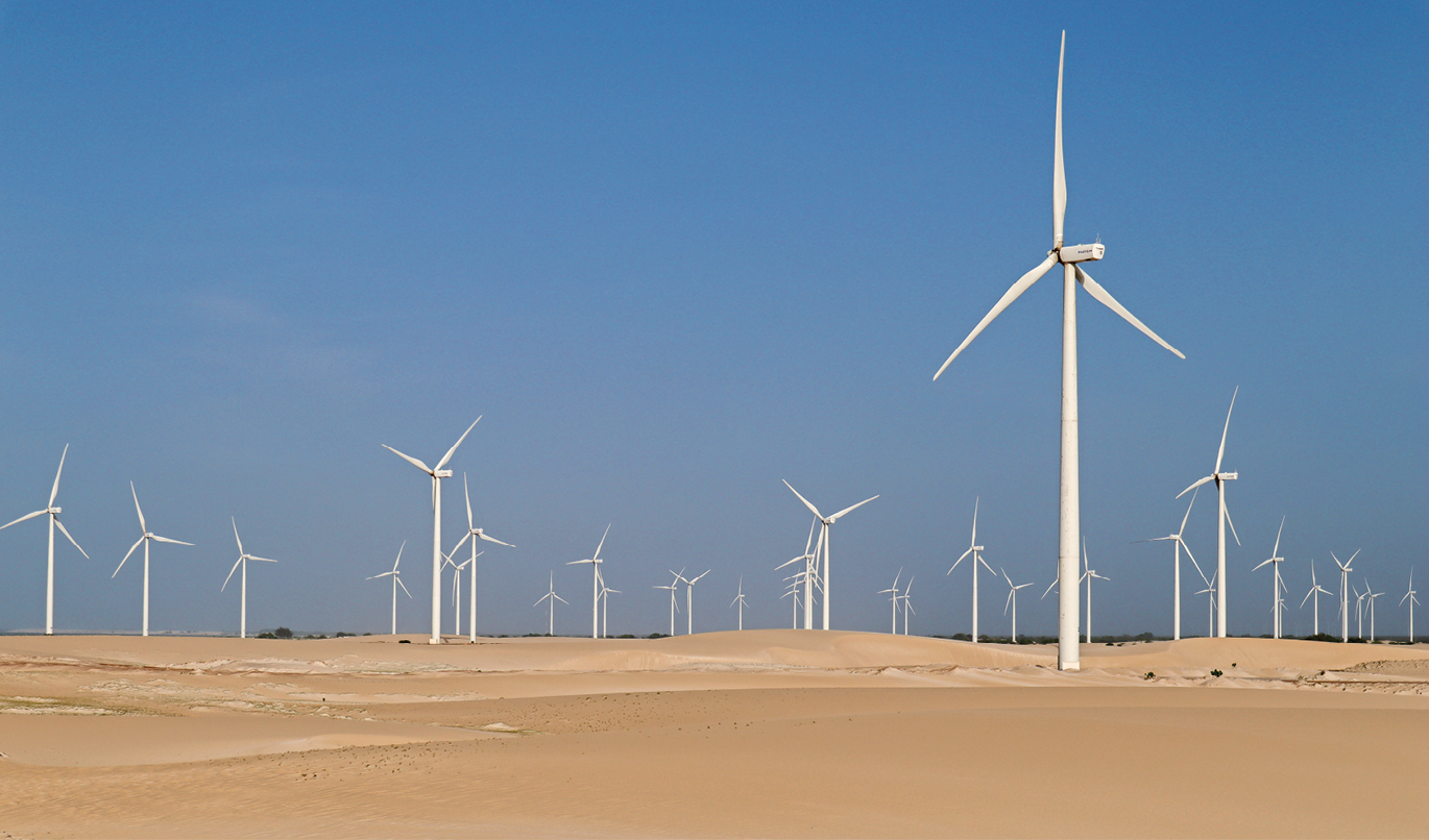 Fotografia. Área com torres de captação de energia eólica. Cada uma é formada por uma haste e três hélices. O chão é de terra. O dia está ensolarado e sem nuvens.