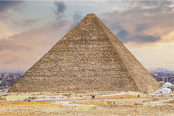 Fotografia: Pirâmide antiga no deserto. Ao fundo, prédios e construções de uma cidade.