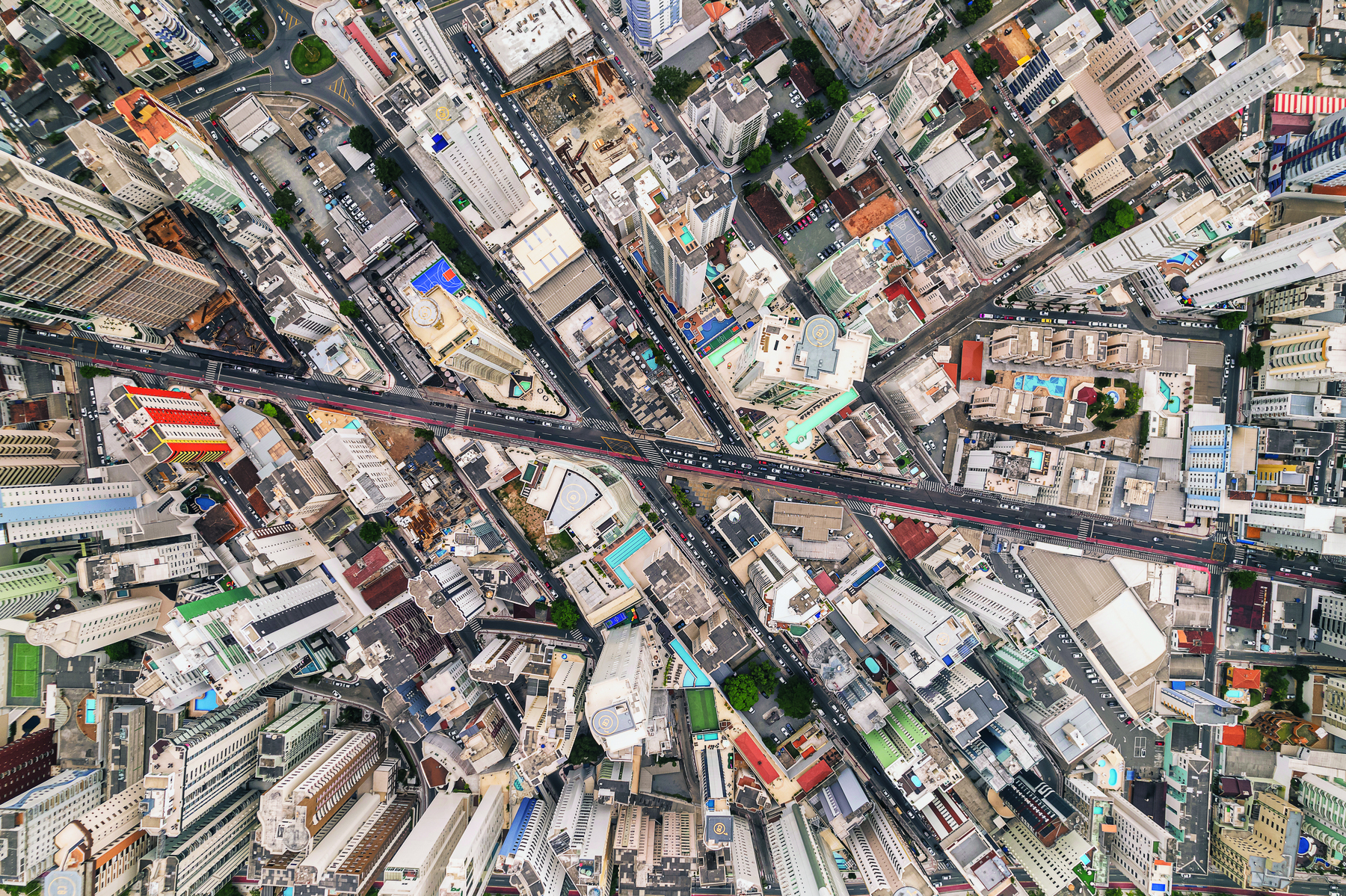 Fotografia. Vista aérea de uma região de uma cidade. No centro, na horizontal, avenida com veículos. Em volta, outras ruas e muitas construções, como prédios e casa.