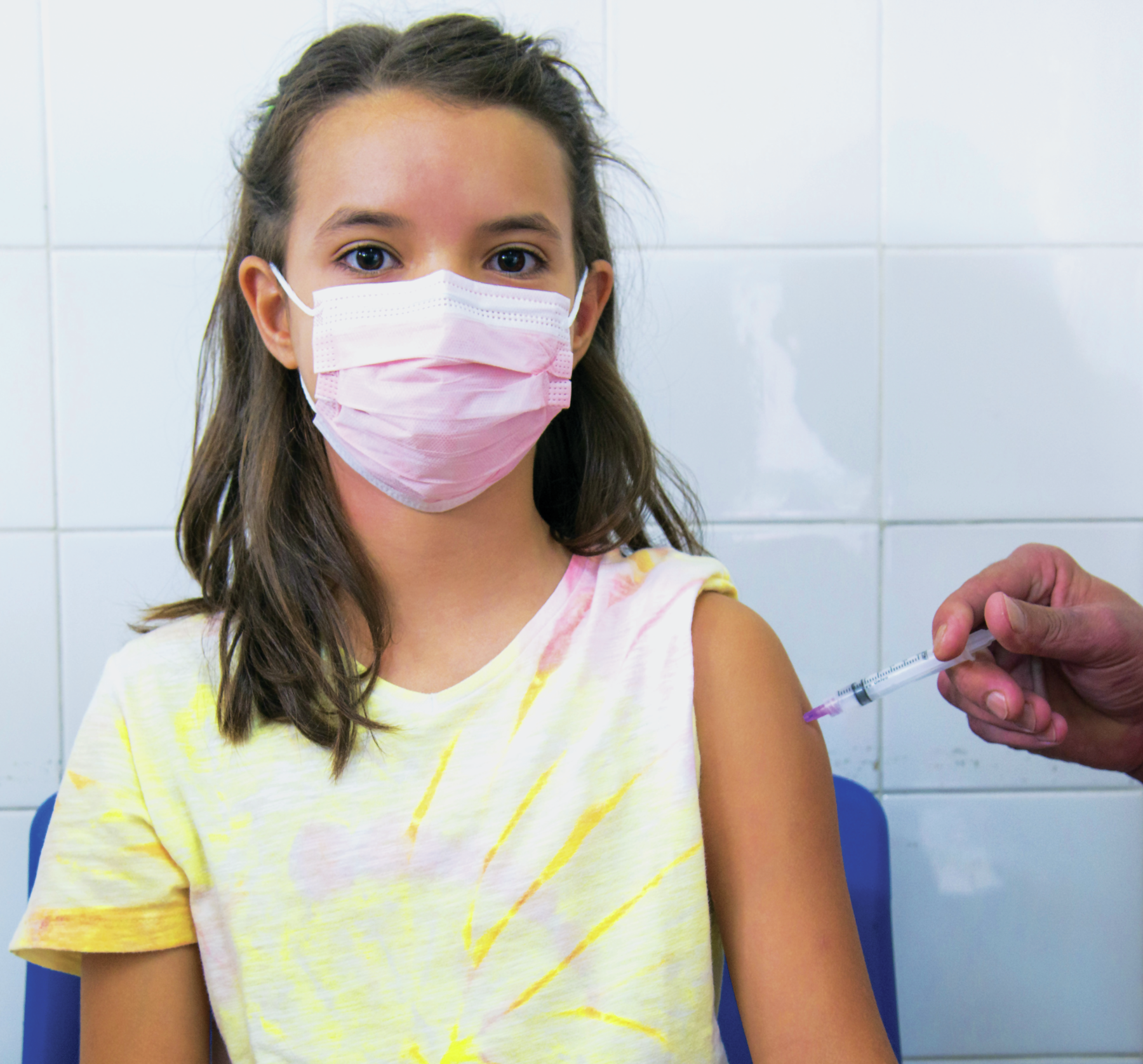 Fotografia. Menina branca, cabelo castanho, vestindo máscara rosa e camiseta amarela. Está com a manga do blusa do lado esquerdo levantada, com uma injeção sendo aplicada por uma mão negra.