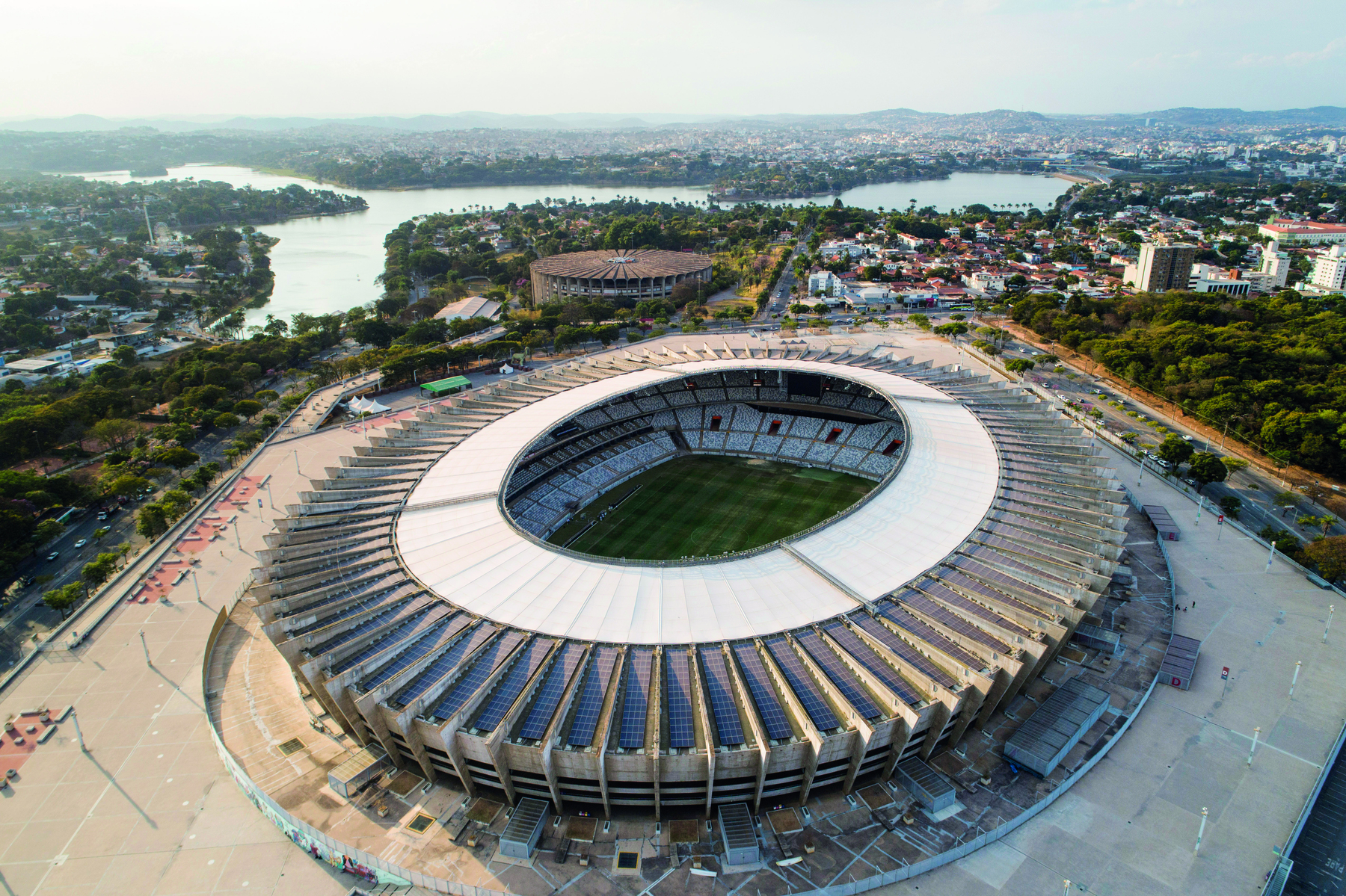 Fotografia. Vista aérea de estádio de futebol. Possui cobertura cinza na parte da arquibancada, circular. É possível observar parte do campo e da arquibancada. Ao fundo, cidade.