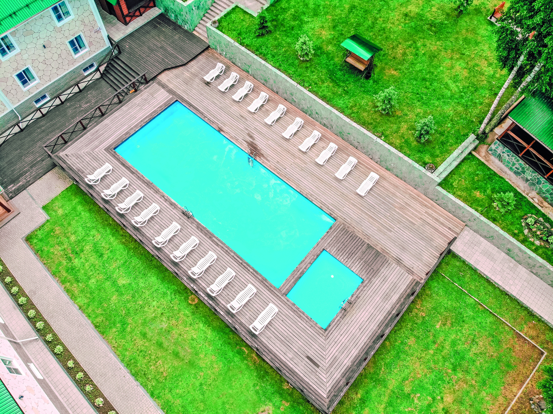 Fotografia. Um piscina superfície retangular maior com água e outra menor, ao lado. Ao redor, espreguiçadeiras na cor branca. Em volta, gramado e outras construções.