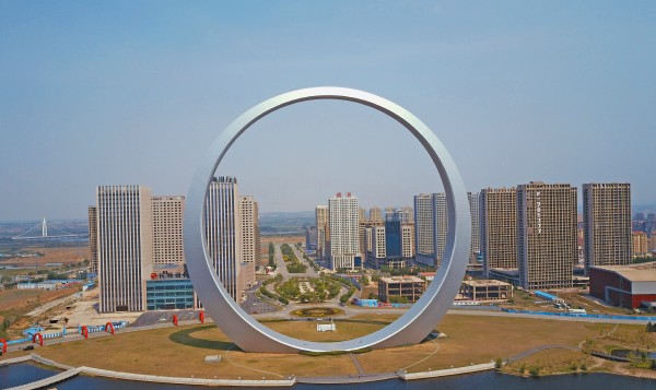 Fotografia. Escultura que se parece com uma circunferência. A escultura está apoiada em um gramado. Ao fundo há vários prédios, construções e vias. O céu está azul e sem nuvens.