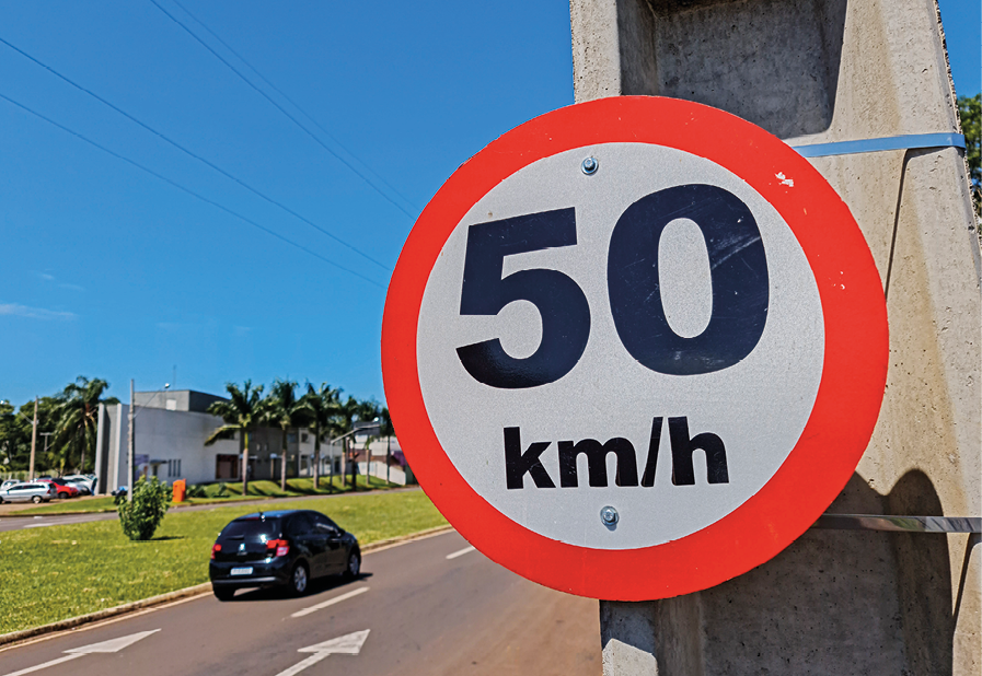 Fotografia. Avenida asfaltada com um carro preto passando. Ao fundo, gramado com construção. À frente, pendurada em um poste, placa branca em formato de círculo com borda vermelha e texto em preto, no centro: 50 quilômetros por hora.