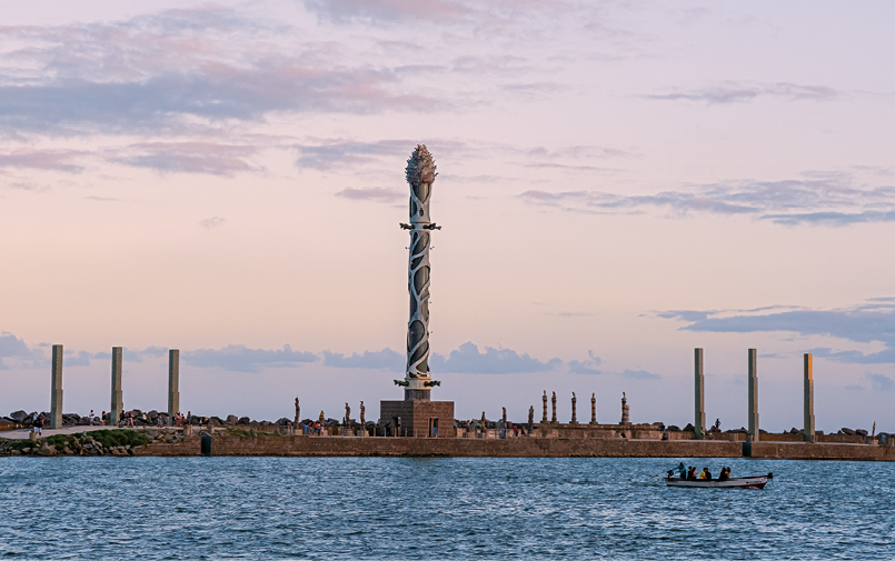 Fotografia. Primeira imagem. Mar com uma área construída. Há várias esculturas verticais e, ao centro, uma escultura vertical maior, que chega próxima à borda de cima da fotografia. Ao fundo, céu.