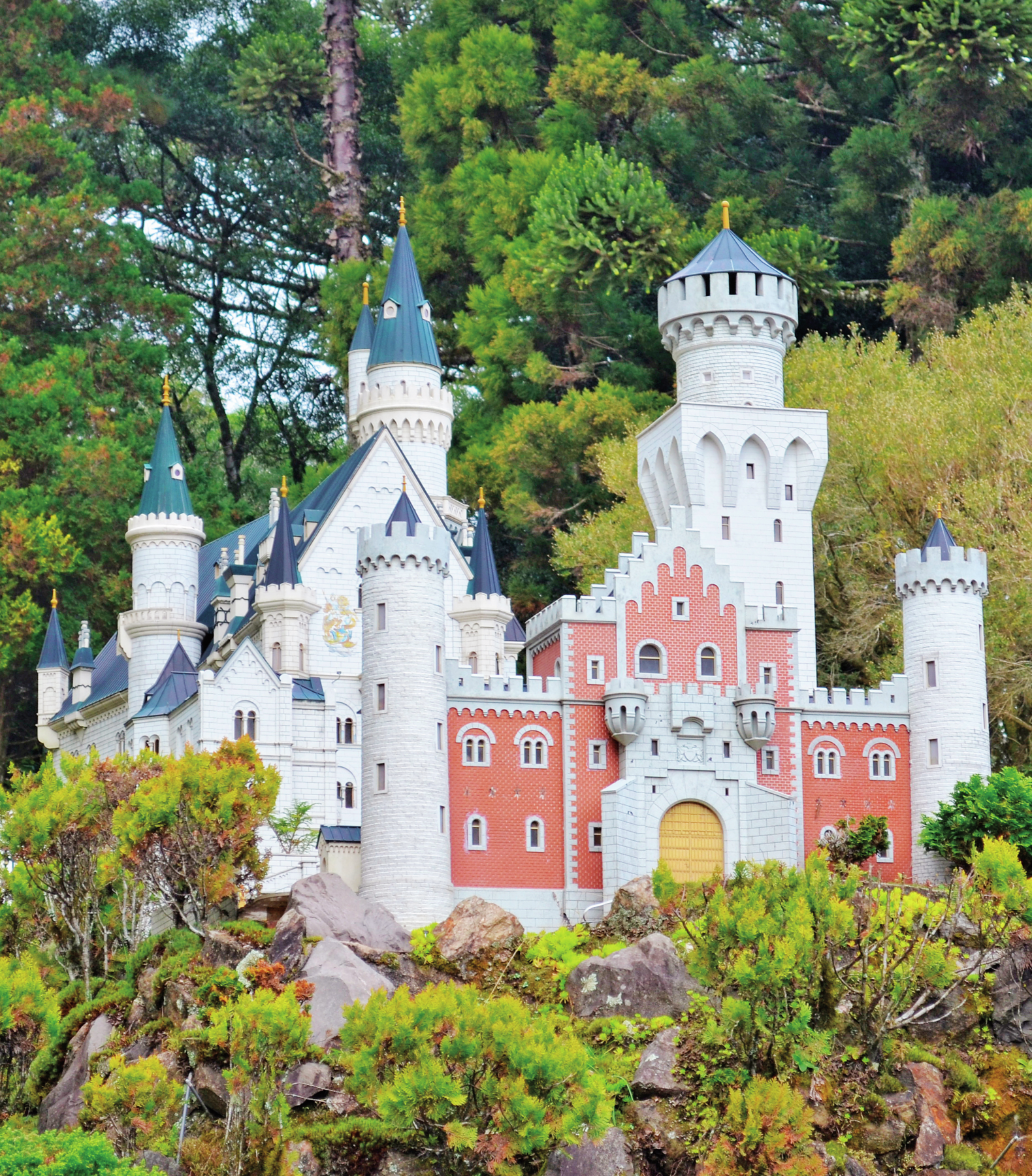 Fotografia. Miniatura de castelo cinza e branco, com fachada na cor vermelha e porta na cor amarela. Possui torres com telhados em formato de cone na cor verde clara. Na parte inferior, árvores de vários tipos e tamanhos. Ao fundo, vegetação.