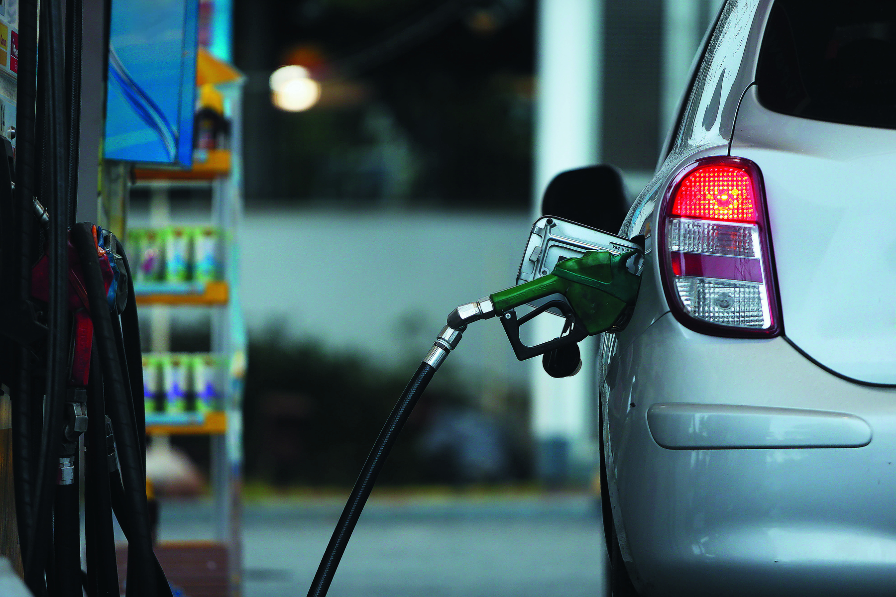 Fotografia. Traseira esquerda de um veículo prata, com farol traseiro vermelho aceso. Está parado em um posto de gasolina, sendo abastecido.