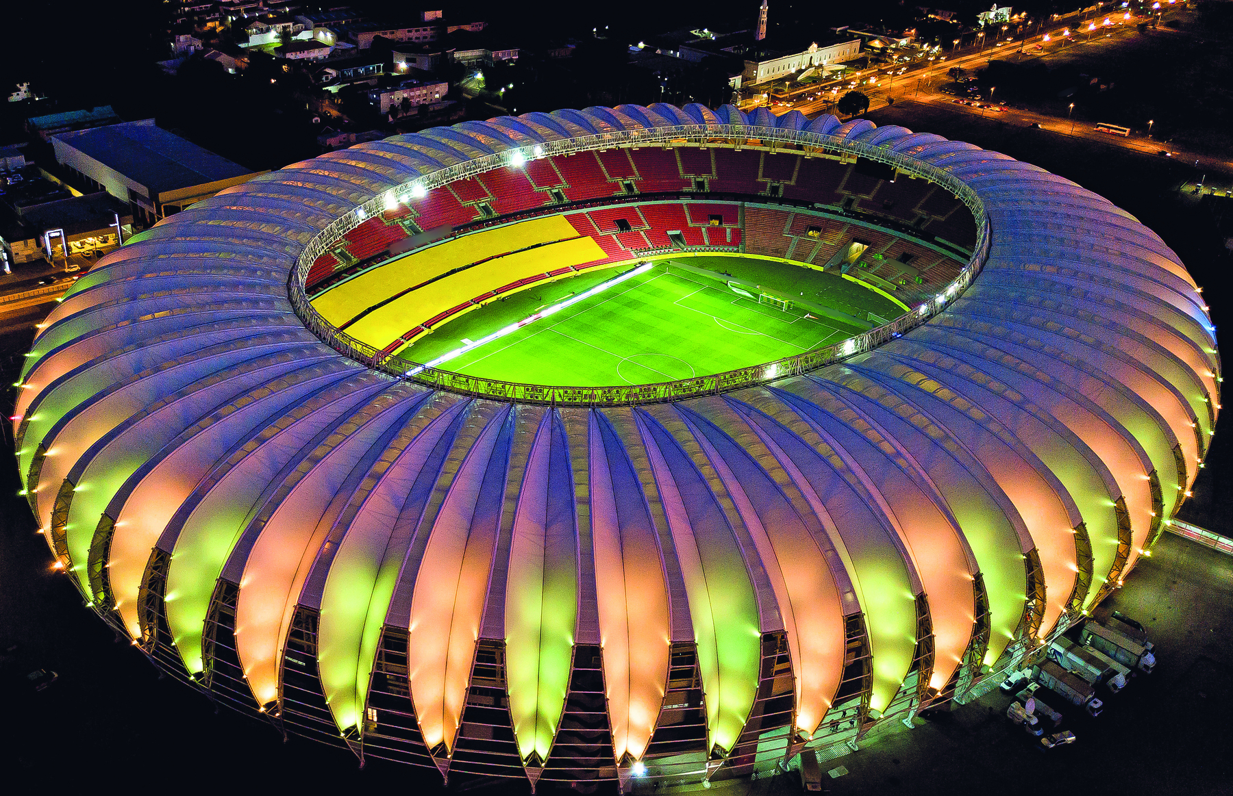 Fotografia. Vista aérea noturna de estádio de futebol. Possui cobertura cinza na parte da arquibancada, circular, e está iluminada na cores verde e rosa. É possível observar parte do campo e da arquibancada vazia, que tem as cores amarela e vermelha. Ao fundo, cidade iluminada.